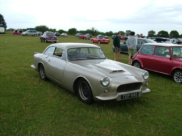 Fibreglass 2+2 classics - Page 1 - Classic Cars and Yesterday's Heroes - PistonHeads - The image showcases a classic car show with a large silver classic sports car parked on bright green grass, adjacent to a vintage red car. In the background, there are other cars including a classic grey convertible and a blue convertible. A group of people, one of whom is holding a camera, are standing behind the cars, admiring them. The sky is overcast, creating a serene atmosphere.