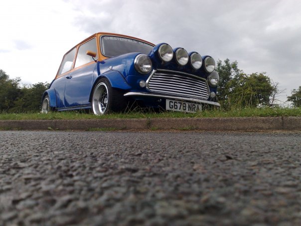 my little mini - Page 1 - Readers' Cars - PistonHeads - The image features a vintage blue Mini Cooper parked on the side of a road. The Mini Cooper's car body is painted blue and it sports a white license plate. The car is curved and has distinctive, clear orange headlights. The vehicle is positioned in the corner of the street, possibly on the edge of a grassy field, with a blurred background. The lighting suggests it might be an overcast day.