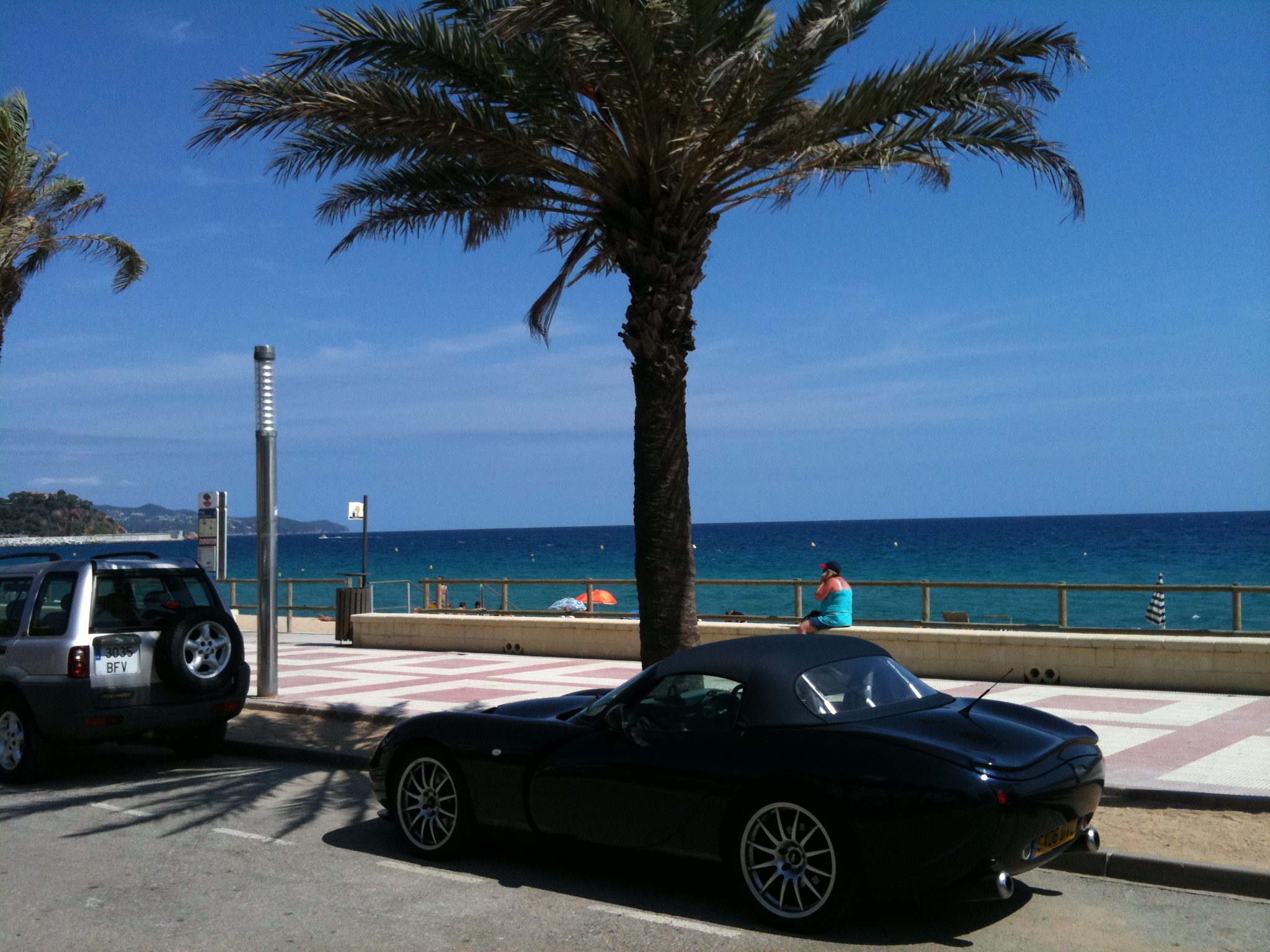 Ultimate Car Spotter Thread - Page 4 - General Gassing - PistonHeads - The image captures a serene beachfront scene. A black sports car is parked on the side of the road, facing the ocean, appearing like a silent observer of the beauty around it. A palm tree stands tall nearby, adding a touch of tropical vibe. The road is situated right next to a boardwalk with a metallic fence and a few umbrellas scattered around, suggesting a popular spot for locals and tourists alike to take in the view. The vast expanse of the ocean stretches into the horizon, complementing the clear blue sky that adds to the overall tranquility of the place.