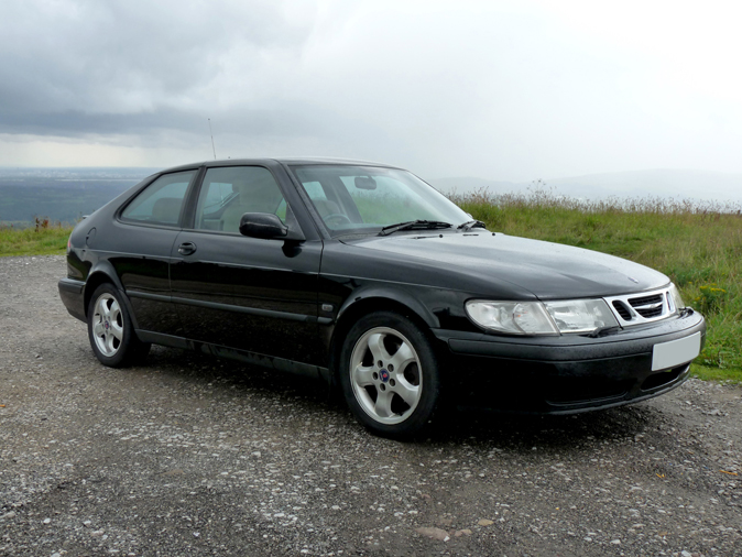 The image features a dark-colored compact sedan parked on what appears to be a combination of paved and gravelled area. The car's body and wheel rims are black, with the vehicle positioned in a dynamic angle that provides a side view of the car. The sky is overcast, and the horizon is visible with a mountainous landscape beyond. There is a single distinguishing feature in the grill of the car.