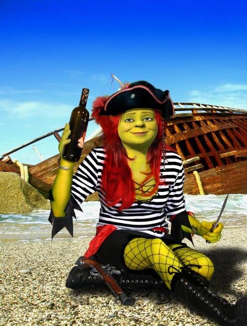 Scullybbb - The image shows a person with green skin sitting on a rocky beach under a clear blue sky. They are wearing a black and white striped pirate shirt, yellow crochet fingerless gloves, and a pirate hat topping a red wig with curly bangs. The individual is holding a green bottle in one hand and a knife in the other. Behind them, there's a rusted shipwreck with wooden planks and ropes.