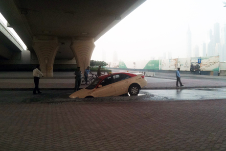 Why do they sell water soluble cars in the Middle East? - Page 1 - Middle East - PistonHeads - In the image, a beige car, seemingly upside down, is prominently featured near the edge of a flooded street. A group of people are standing near the car, possibly discussing how it got into its current position. The car blockage is creating a small pool of water, highlighting both the unusualness of the scene and the severity of the flooding. The overcast sky adds a gloomy atmosphere to the scene, suggesting possible weather-related disasters. The distance shows skyscrapers in the vicinity, potentially hinting at an urban environment.