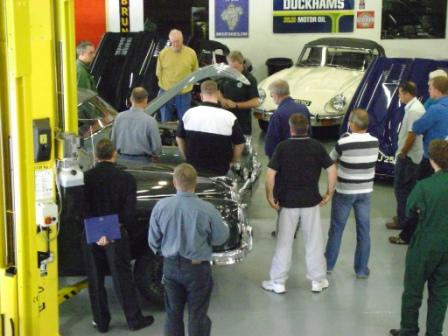 Pictures from Paul Roach's Open Day - Page 1 - Classic Cars and Yesterday's Heroes - PistonHeads - The image captures a social gathering around vintage cars. In the foreground, a group of men in casual attire are admiring and discussing a restored silver car. Further in the background, more individuals are observing the one man on the left who is sitting alone in a white antique vehicle. 

The setting appears to be an indoor garage or showroom, possibly a classic car exhibition as suggested by the overall ambiance. Various posters adorn the walls, which might be related to the event or event sponsors. The presence of these signs and the crowd of people suggest that this event is likely part of a wider exhibition dedicated to classic and antique vehicles. 

The large dining table in the background further hints at the possibility of the event featuring discussions or showcasing related products and memorabilia. In summary, the image portrays a convivial scene of classic car enthusiasts gathered for a shared interest in vintage automobiles.