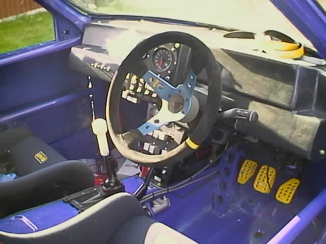 MG Metro 6R4- Under The Plastic - Page 1 - Classic Cars and Yesterday's Heroes - PistonHeads - The image displays the interior of a sports car with a blue driver's seat. The steering wheel is attached to the dashboard, showcasing a variety of gauges and controls. The steering wheel has clamp straps around it, possibly for safety purposes. In the foreground and floor area, there is a blue ejection seat. The car's window curves subtly towards the back, typical of sports cars for structural integrity. The overall scene suggests a preparation for a drive or an exhibition of the interior.