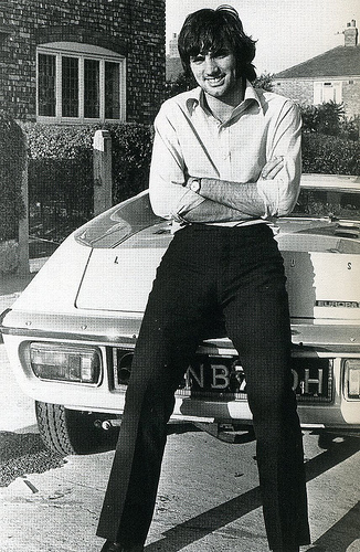 Pistonheads Classics Famous - The image appears to be an old photograph, showcasing a man seated on the hood of an older white car. He is smiling and wearing a white shirt and black trousers. The car is parked in front of a building, and there's greenery visible in the background. The style of the clothing, the design of the car, and the quality of the image suggest it might be from a few decades ago.