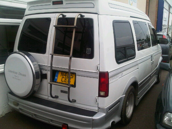 Astro Vauxhall Fails Chevrolet Pistonheads Combi Spot - The image shows the back of a white, long-wheelbase van parked on the street. Notably, there is a mounted bicycle rack on the back, which is empty. The van is situated next to the curb, adjacent to a building with a window that can be seen in the background. The van appears to be in standard condition, indicating it's likely part of a commercial or service fleet due to its size and purpose. There is no specific detail about the license plate, although it's note-worthy that the registation is written in a foreign alphabet, possibly an East Asian script.