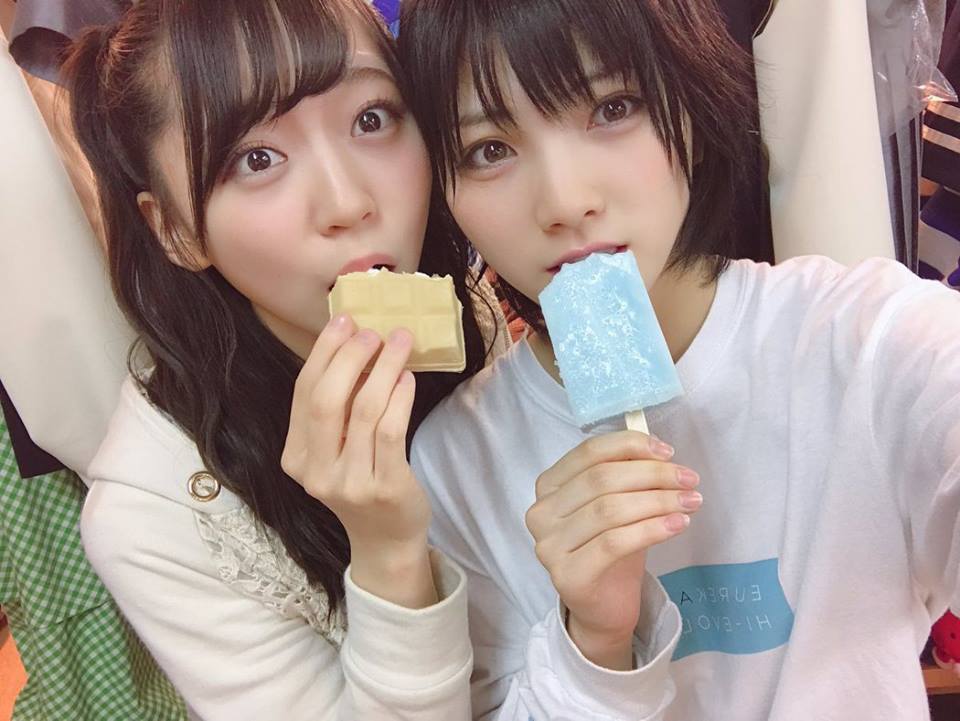 This image captures a sweet moment between two women sharing ice cream. Both women have medium-length dark hair and are looking into the camera. They are biting into their ice creams but appear to be taking a break from eating, as the ice creams are visibly intact. The colored clothing suggests a casual setting, and their smiles and the act of sharing ice cream convey a sense of happiness and friendship.