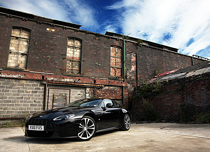 Who has a V12 Vantage? - Page 1 - Aston Martin - PistonHeads - The image features a sleek, black sports car parked next to an old brick building. The car is likely an Audi models, as indicated by the vehicle's design language and badge. Its silver rims contrast nicely with the black body, suggesting a focus on both aesthetics and practicality. The building has a rugged charm with its red brick facade, and there's a missing window and cracked bricks, indicating a touch of neglect. The massive wrought iron gates add an element of grandeur to the scene. All of this is set against a backdrop of a cloudy sky, giving the picture an overall moody and atmospheric feel.