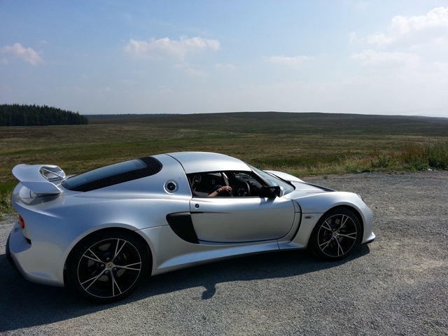 lets see your Lotus(s)! - Page 1 - General Lotus Stuff - PistonHeads - The image showcases an **AdvancED V12 GT** sports car parked on a **gravel road** near a **grass field**. The car is facing us, with the **windshield** open, its **gleaming silver body** reflecting the surrounding area. A person, presumably the driver, is visible in the car, enjoying the serene environment. The backdrop of the scene is dominated by a clear **blue sky** and rolling **green hills**.