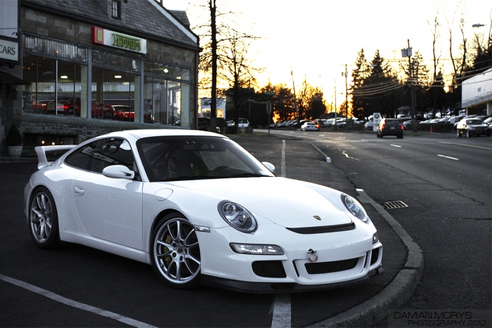 Pistonheads - This is a black and white photograph of a white Porsche sports car parked on the side of a road. It's positioned in front of a building with striped awnings. The car is facing to the left and the setting appears to be during either sunrise or sunset, given the warm and soft light illuminating the scene. The road has multiple lanes and another car is captured in the background moving away from the viewer.