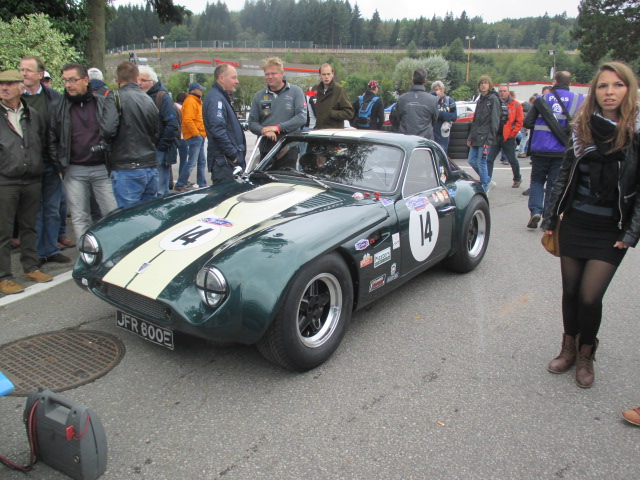 Spa 6 hours, 2015 - Page 1 - Classics - PistonHeads - The image showcases a vintage sportscar parked by the side of a road, drawing the attention of onlookers. The car, predominantly green with a white stripe down the middle, is well-maintained with the door open, dressed up with various stickers. It's hard to miss the small buoy-shaped object next to the car. Several people are around the car, some posing for the camera and others examining the vehicle, all seemingly captivated by the car's classic charm.