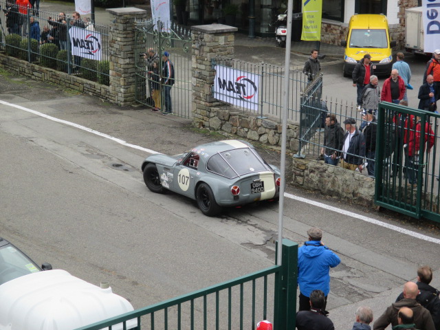 Spa 6 hours, 2015 - Page 1 - Classics - PistonHeads - The image shows a scene where a gray-green car is driving down a city street. Spectators are present on the sidewalk, watching the car from behind a fence. Near the car, there's a white board that says "DIET", indicating that this could be an event or demonstration. In the background, there is a fence, a flag, a building, and a yellow car. The image captures a moment of curiosity and interest from the onlookers towards the passing car.