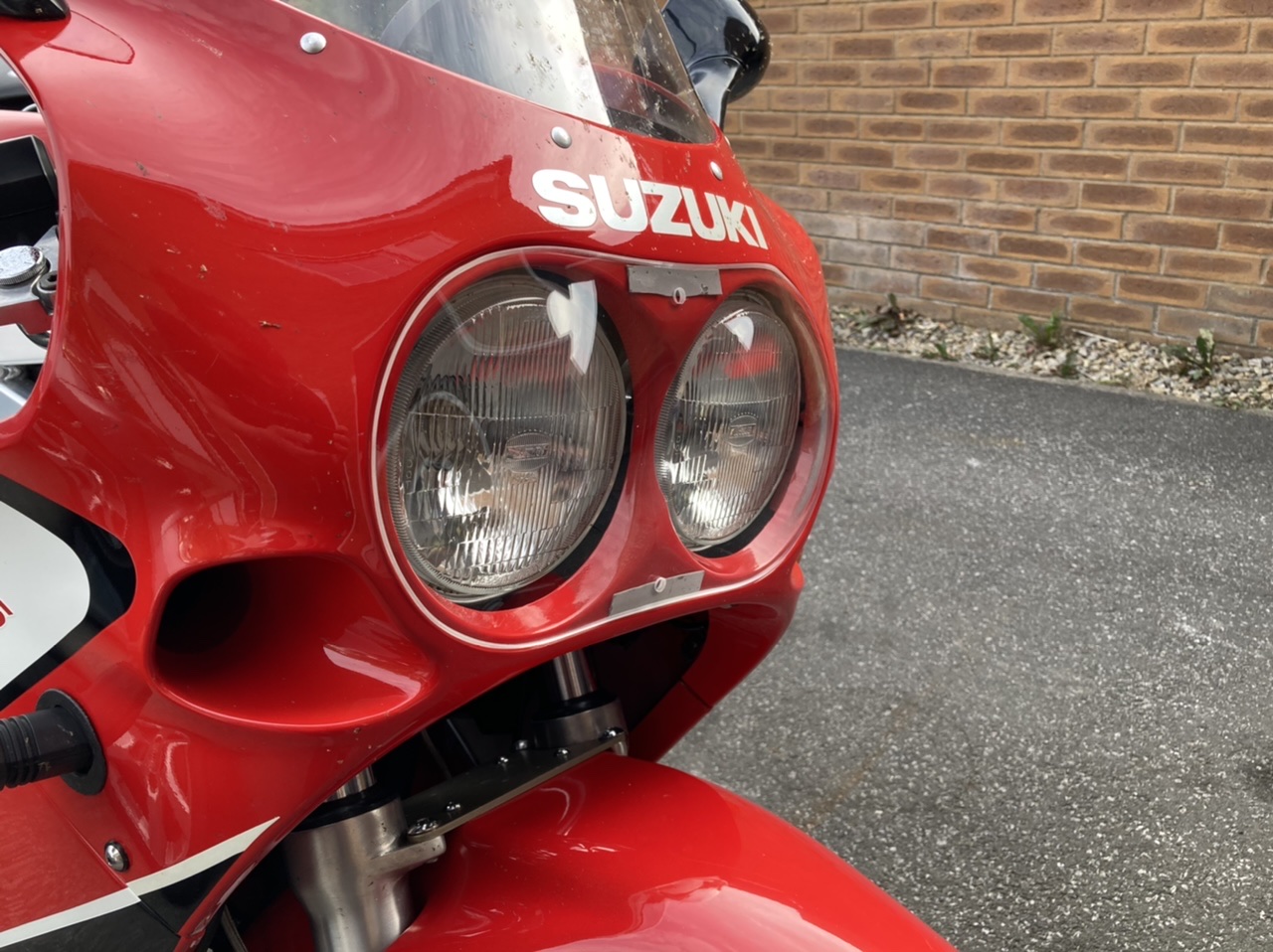 The Tasteful Modification Thread  - Page 5 - Biker Banter - PistonHeads UK - The image shows a close-up of the front of a red motorcycle, featuring a prominently displayed logo that reads "SUZUKI" along with other branding. The bike has dual round headlights and is parked on what appears to be a driveway with brickwork in the background. The style of the image suggests it may have been taken to showcase the design or branding of the motorcycle, possibly for marketing or catalog purposes.