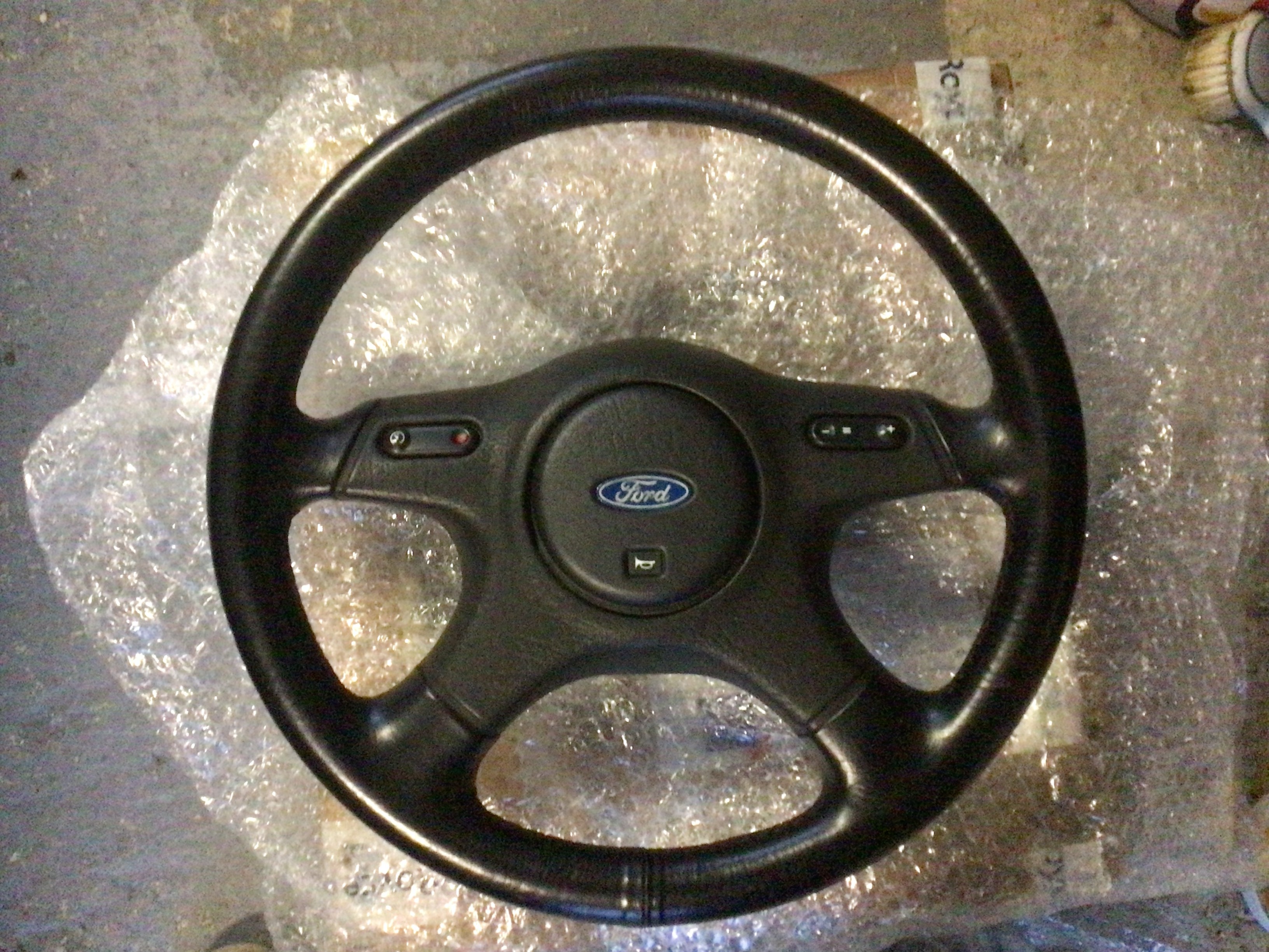 Ford Granada - Attainable 90s Interesting Ford - Page 2 - Readers' Cars - PistonHeads UK - The image shows a black steering wheel with a silver center and the Ford logo. It is placed inside a transparent protective bag, which is situated on a surface that appears to be carpeted. The car's dashboard or a similar part of the vehicle is visible around the steering wheel. In the background, there's a glimpse of another object wrapped in plastic, suggesting this might be a storage area for car parts.