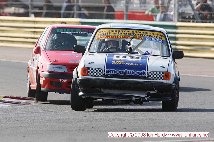 Pistonheads - This image captures an exhilarating moment on a racetrack where two cars, with numbers "88" and "99" on the front, are engaged in competition. The first car, a red sports car, is overtaking the second car, a white car, as they race around a right-hand corner. Both cars are maintaining a tight line on the track, highlighting their skilled drivers. A track worker can be seen walking across the stands in the background, adding a dynamic element to the scene.