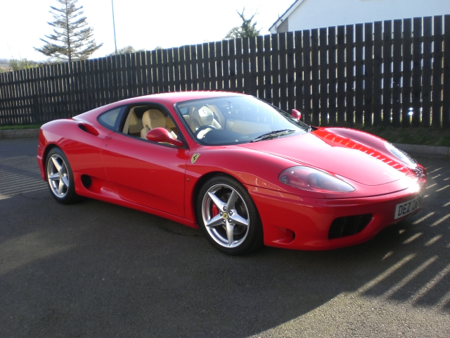 The image portrays a vibrant red convertible sports car parked in a sunlit lot, smiling out under the clear blue sky. The car is positioned facing us, showcasing its sleek design and alloy rims. The wooden fence in the background provides a rustic contrast to the high-end sophistication of the vehicle. The scene is bathed in the hues of a warm summer afternoon.