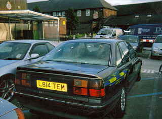 RE: Rover 825 Sterling: Shed of the Week - Page 1 - General Gassing - PistonHeads - The image shows a scene of a parking lot with several cars parked. A distinctly painted hatchback car with a yellow license plate reading "LB14TEM" is prominently featured in the foreground on its side. The background includes a brick building and some vehicles in the distance, but the focus is on the parked car with a license plate that gives it some character.