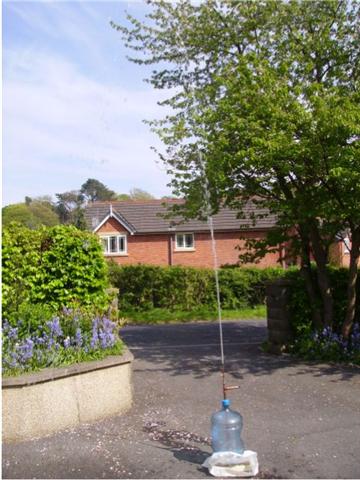 A fire hydrant in front of a brick building - Pistonheads - The image showcases an outdoor scene with several elements. A large, clear plastic water bottle is precariously tipped over and spewing water onto the ground. The bottle is on a driveway that leads to a brick building with a pitched roof. The building appears to be a house and is surrounded by a well-maintained garden. There's a tree prominently in the foreground, adding a touch of nature to the scene. The overall atmosphere appears to be a quiet suburban setting.