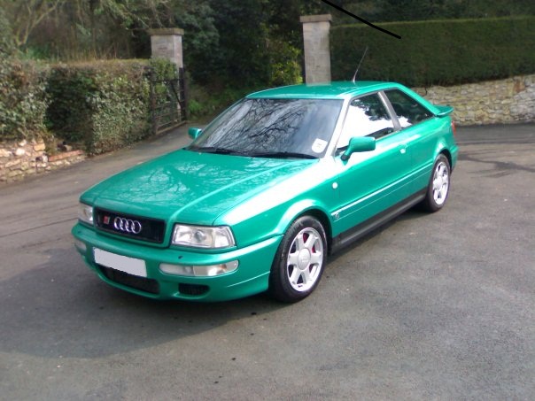 Audi S2, any good? - Page 1 - Audi, VW, Seat & Skoda - PistonHeads - The image depicts a vibrant green Audi Quantum parked on a concrete surface. It's equipped with red and black accents that accent markings on the side of the car, and it sports white wheels with a multi-spoke design. The car is positioned at an angle that shows both the front and rear, allowing for a clear view of its sleek exterior. The background reveals a verdant hedge and a metal fence, suggesting a private property or an upscale parking area.