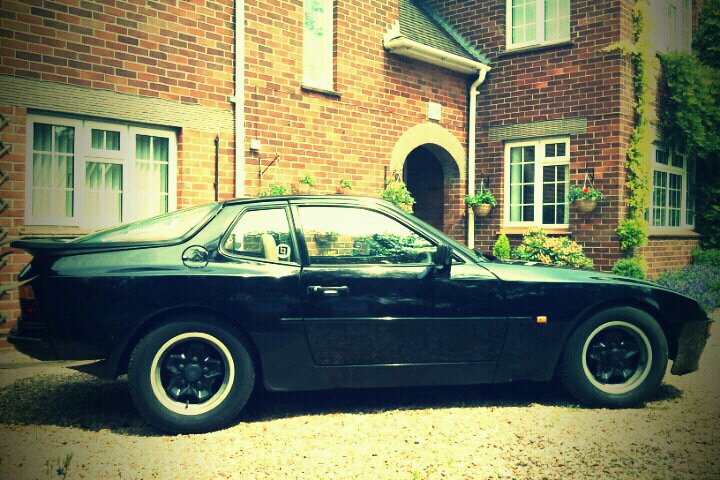 Hate Love Pistonheads - The image showcases a classic black sports car parked in front of a red brick house, displaying its hood and showing the overall front view. The car is positioned on the driveway, adjacent to the front door of the house. The house features a neatly trimmed green hedge and plant pots, with windows that add architectural detail to the scene. There's a tree visible in the background, suggesting a suburban setting. The car is spotted, which enhances its aesthetics and makes it stand out.