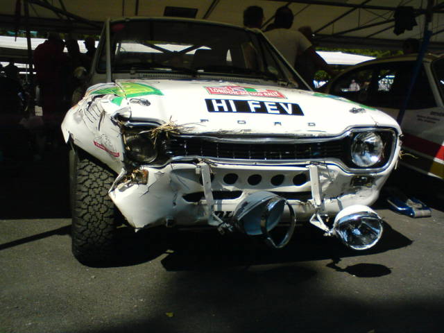 Ford Fev Rip Escort Fos Pistonheads Goodwood - The image shows a frontal view of a classic car with a predominantly white body. The car's hood is deformed, and the left headlamp is missing. The vehicle is surrounded by a group of people who appear to be standing at an outdoor location, possibly a garage or exhibition space. The setting is not completely clear, but it looks like the car is being viewed or evaluated in a social context.