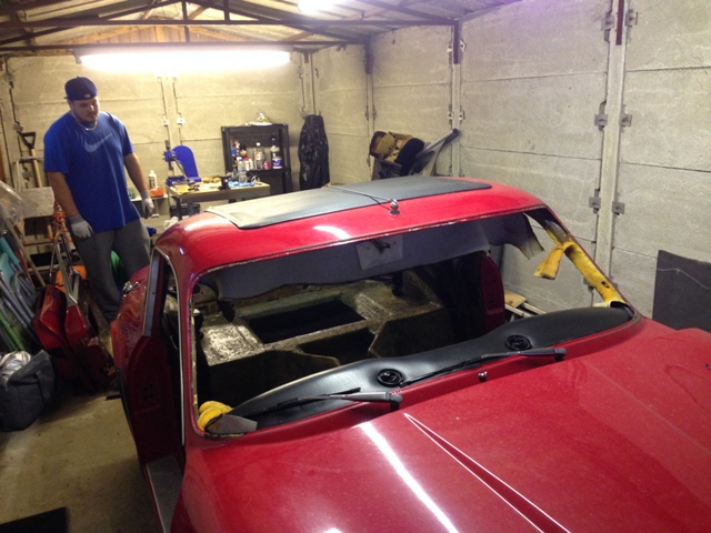 A man is standing in front of a parked truck - Pistonheads - The image features a man in a blue shirt standing near a red sports car. The man is operating a power wrench near the top of the car's door frame. The car's top is missing, revealing its metal framework. The setting appears to be a garage, with various tools and equipment scattered around. There is no visible text in the image.