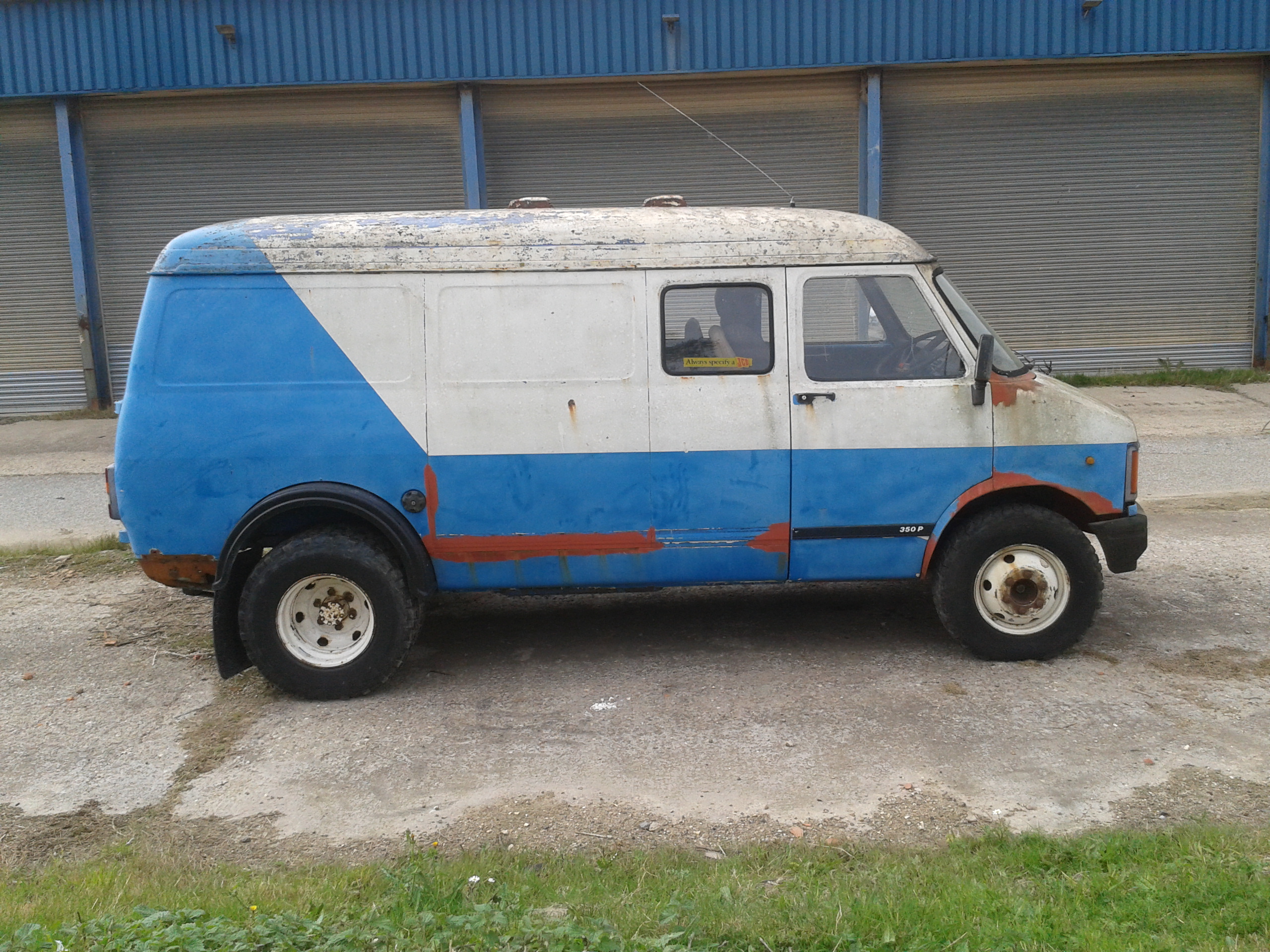 why no love for beford/vauxhall? - Page 3 - Classic Cars and Yesterday's Heroes - PistonHeads - The image shows a traditional van parked on a paved surface. The van is blue and white with the back painted in red and rust streaks visible on the side. The design on the side includes a retro style and appears to be an old model, possibly a utility van. The background includes a garage with large blue doors, suggesting that the van is parked in a driveway or entrance of a commercial or residential building. The van has a spare tire mounted on the rear, which is a common feature for older vehicles and vans designed for utilitarian purposes.