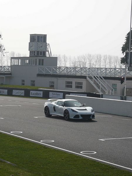 At Goodwood today..... - Page 1 - Goodwood Events - PistonHeads - The image shows a high-performance sports car, specifically a Lamborghini, in motion on a track. The vehicle is white with black accents, and it appears to be traveling at a fast speed. The setting of the track is indicated by the presence of a white building with a clock tower in the background, which is presumably a part of the race track's administrative complex. The sky is overcast, suggesting an overcast day. The track itself is well-maintained with clear markings, and the overall atmosphere of the image is dynamic and focused.