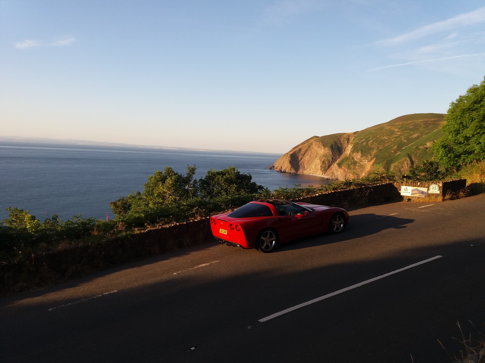 The £7700 Corvette C6 - Page 10 - Readers' Cars - PistonHeads - The image captures a vibrant scene, featuring a red sports car parked on a twisty road that overlooks a vast body of water, possibly an ocean or a large lake. The car, with its sleek design and imposing tires, is positioned near the viewer, with its interior detail clearly visible through the open top. The road is marked with white lines, indicating paths of travel or perhaps parking restrictions. Beyond the car and the immediate surroundings, the landscape opens up to reveal a rugged coastline and distant hills, creating a dramatic contrast between the natural environment and the modern vehicle. The perspective of the photo suggests it was taken from a distance, looking back towards the car, which adds a sense of nostalgia to the scene.