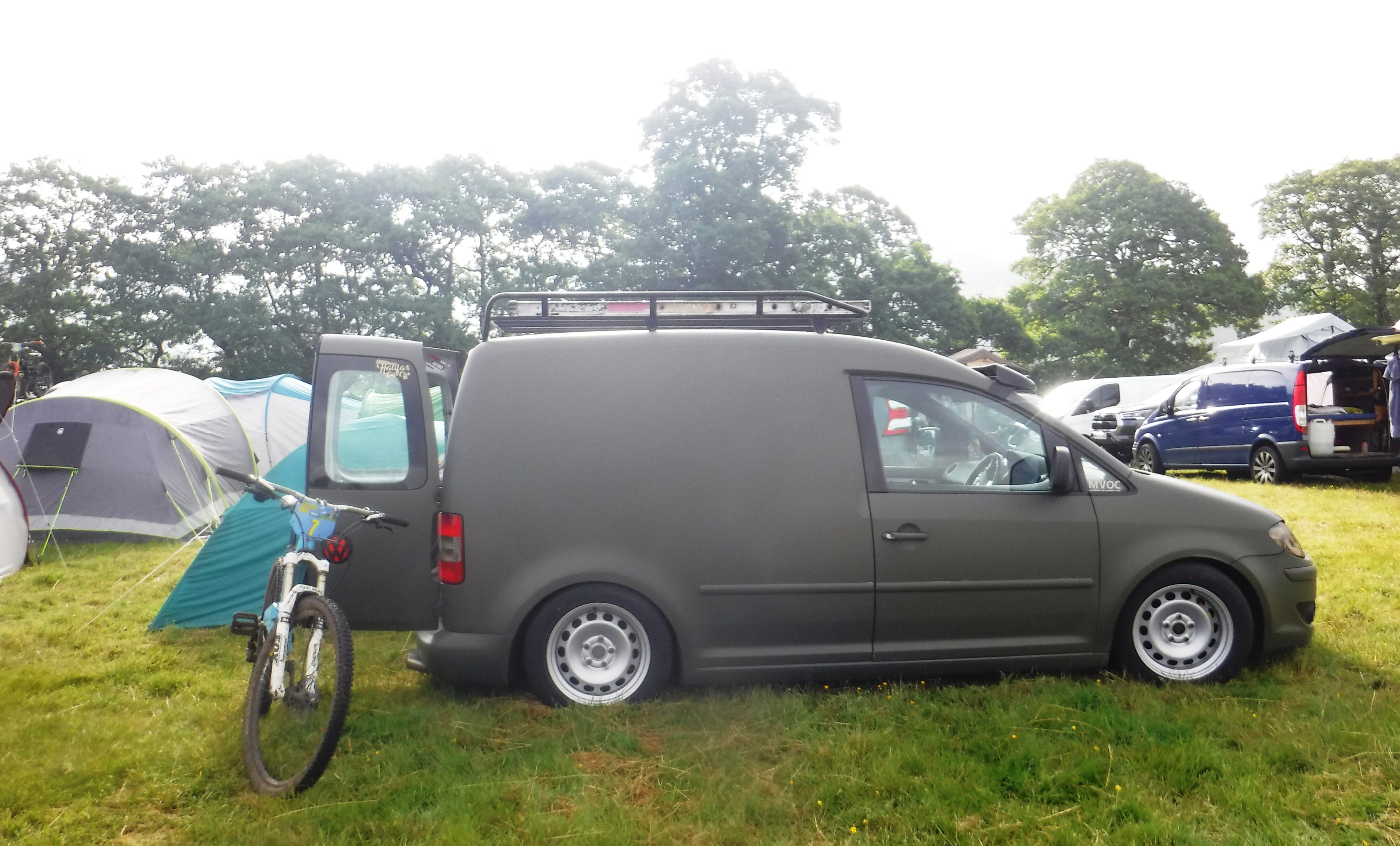 Show us your transport vehicle. - Page 9 - Pedal Powered - PistonHeads UK - The image depicts a scene from an outdoor event, likely a camping or festival setting. There's a black van parked in the middle of the frame with its doors open, revealing items inside. A bicycle is leaning against the side of the van, suggesting someone has momentarily left it unattended. In the background, there are other vehicles and tents, indicating that this location is part of a larger gathering or campsite. The setting seems to be an open field or park with grass visible under the parked vehicles.