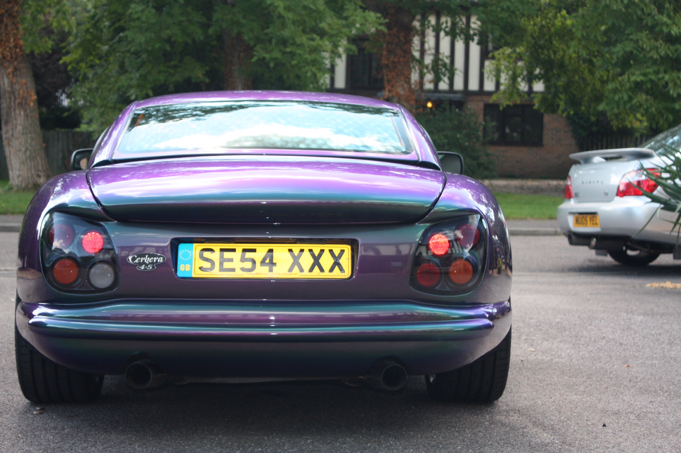 Nice Ass - Page 3 - Tamora, T350 & Sagaris - PistonHeads - This image features a purple sports car parked on a street. The car is facing to the right, with the back windshield visible. A distinctive license plate is affixed to the back, reading "SE54 XXX". The car has a striking purple color with a glossy finish, reflecting the light in the surroundings. In the background, there appears to be a residential area with trees and a building visible. Another car is partially visible to the right in the image.