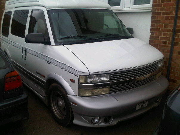 Pistonheads Chevrolet Fails Astro Combi Vauxhall Spot - This image features a clean, white rocky mountain 4-wheel drive parked on the street. The van is sleek and modern, with its chrome details adding to its distinctive style. Its wheels appear to be in good condition, and it has a fresh, clean look. The background is a brick wall, with a bunch of twigs scattered along the ground. The setting suggests a suburban area, and the visibility of the license plate identifies it as a west coast van.