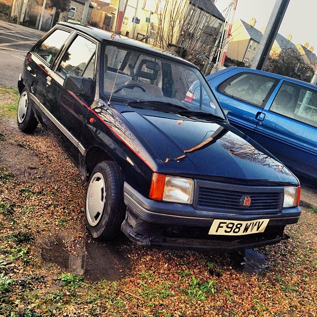 A car parked on the side of the road - Pistonheads - The image features a dark blue hatchback car parked on gravel next to a blue station wagon. The car has a distinctive bulge in the front hood, which is characteristic of some classic vehicle models. The gravel surface appears to be well-trodden, indicating frequent use or possibly sparse parking opportunities. The environment suggests a residential or suburban area with buildings visible in the background. The sky is overcast, and the lighting is soft and diffused, suggesting either an early morning or late afternoon scenario.