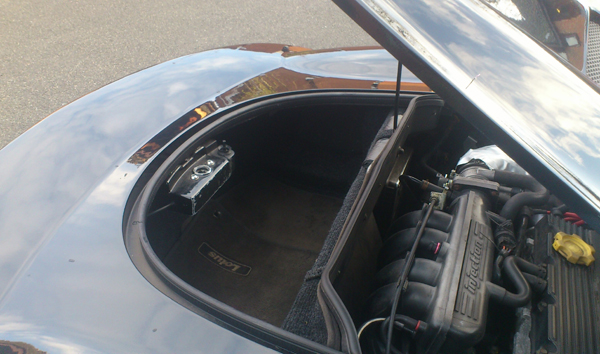 Pics of your bootspace pls - Page 1 - Elise/Exige/Europa/340R - PistonHeads - The image captures a scene from beneath the hood of a car. The car's trunk is open, revealing its black interior. The trunk is secured with a latch, indicating that it's meant for storage. The car's engine lies exposed, reflecting the surrounding light and emphasizing the robustness of the mechanical workings. The car's taillight can be seen in the background, suggesting that this view is from a car moving forward. The backroad and sky overhead hint at a rural or suburban setting.