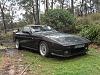 A car parked on the side of a road - Pistonheads - This image depicts a modern, black luxury sports car parked on a grassy area, which appears to be at the edge of a wooded area. The car is facing the viewer, with its sleek design accentuated by the sunlight. There are no visible texts, people, or distinctive brands that are instantly recognizable in the photograph. The photograph's quality and angle suggest it was taken during the day with natural light, and the background, dominated by greenery, implies a calm and serene environment.