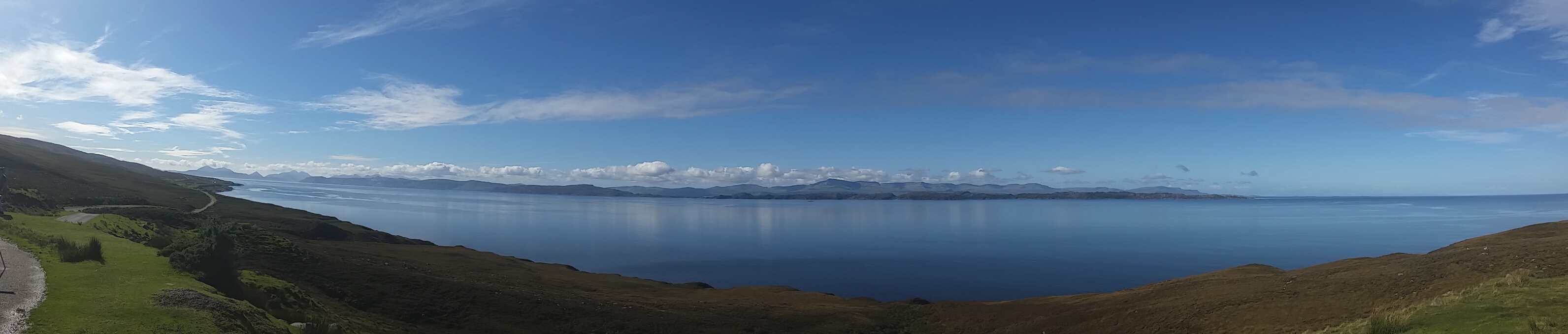 NC500 (north coast road) - Page 13 - Scotland - PistonHeads - The image presents a striking panorama of a landscape dominated by a serene, deep blue body of water, presumably an ocean or a large lake. The water's surface is calm, reflecting the clear blue sky above, dotted with a few fluffy white clouds. On one side of the water, there's a verdant hillside, leading to a road or path that seems to curve around the water's edge. To the right, snow-capped mountains rise majestically against the sky, adding a touch of ruggedness to the otherwise tranquil scene. The image appears to have been captured from a high vantage point, likely from a hill or higher ground, providing a bird's-eye view of this beautiful and serene scenery.