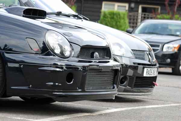 A car is parked on the side of the road - Pistonheads - The image presents a scene from a parking lot where two black cars are parked in front of a building. They are both sleek and shiny, suggesting they are either new or well-maintained. The car in the foreground is an old black Saab with a distinctive grille, while the car in the background is a newer black hatchback with a larger grille and better taillights. The parking lot appears to be near a residential area, as evidenced by the house in the background. The sky is clear, indicating a bright day.