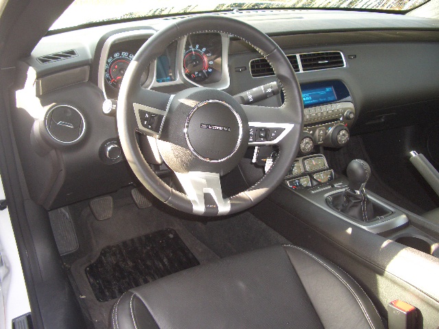 2010 Camaro - Page 1 - Yank Motors - PistonHeads - The image captures the view from the passenger seat of a car, where the black leather seats are visible. The driver's wheel, located at the center of the image, is black with visible buttons and sensors. The dashboard features a control panel with numerous indicators and gauges, and there's a gear shift visible next to the wheel. The general color scheme of the car interior is dark, with predominantly gray, black, and silver tones.