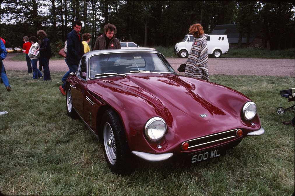 Early TVR Pictures - Page 50 - Classics - PistonHeads - The image captures a scene outside where a classic car, specifically a red BMW with the model number 500 ML, is parked on a grassy area. Five people are gathered around the car, engaged in conversation or preparing for a journey. In the background, there are other vehicles and trees, suggesting a park or a countryside setting. The car's sleek design and the casual attire of the people indicate a relaxed and leisurely atmosphere. This image could represent a group of friends coming together for a road trip in classic vehicles.