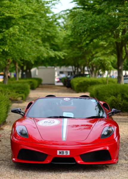 Sunday Supercar Pistonheads - The image depicts a sleek, red Ferrari sports car parked on what appears to be gravel, possibly under a tree given the surrounding greenery. The Ferrari is a two-door convertible model, and it features the iconic Ferrari emblem on its hood and the "573" logo on its side. The gravel path and adjacent trees provide a rustic and natural backdrop to the car, highlighting its design and color against a more subdued environment. The car itself is parked at an angle that allows a view of its front and side, showing an open driver's side door and a hint of its sloped windshield.