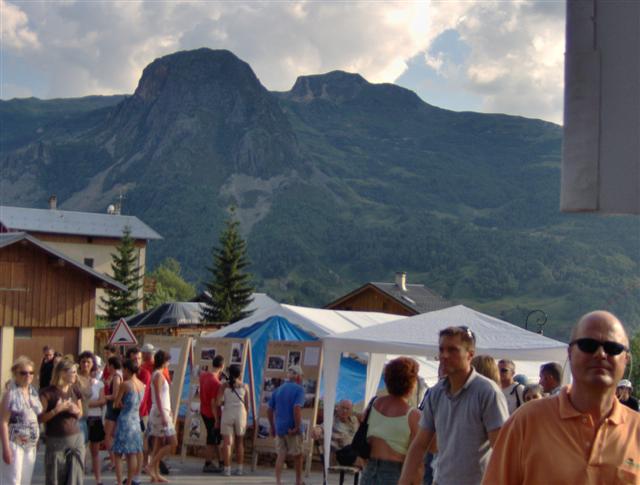 The image showcases a lively scene at an outdoor market. The setting is along a street lined with small shops and stands. A large group of people is gathered around a white tent, which seems to be the center of activity. The crowd includes individuals of various ages and styles, creating a diverse and bustling atmosphere. In the background, a steep mountain looms, providing a dramatic backdrop to the market scene. The presence of early autumn leaves suggests the season, adding a touch of color to the sky. The image captures the vibrancy and liveliness of community gatherings and local markets.