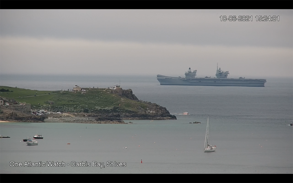 G7 Summit in Cornwall - Page 5 - News, Politics & Economics - PistonHeads UK - The image depicts a coastal scene with two key elements. First, there is a large ship anchored in the water, which appears to be a commercial or cargo vessel due to its size and structure. Second, on land near the water's edge, there are several small boats docked, suggesting this might be a marina or fishing harbor. The shoreline features a rocky coastline with vegetation, and the presence of a small town or settlement can be inferred from the structures visible along the shore.