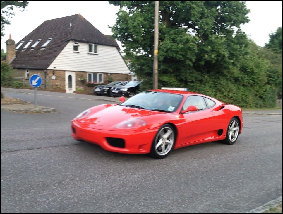 Spotted cars in south coast region [vol. 2] - Page 317 - South Coast - PistonHeads - The image shows a red sports car parked on a residential street. The car is a convertible, characterized by its sleek design and vibrant color. It's seated in front of a blue parking sign. The background features a well-maintained brick house with a roof adorned with tiles. The scene is set during the day under clear skies. The car is positioned on the right side of the road, and there are no other vehicles in sight.