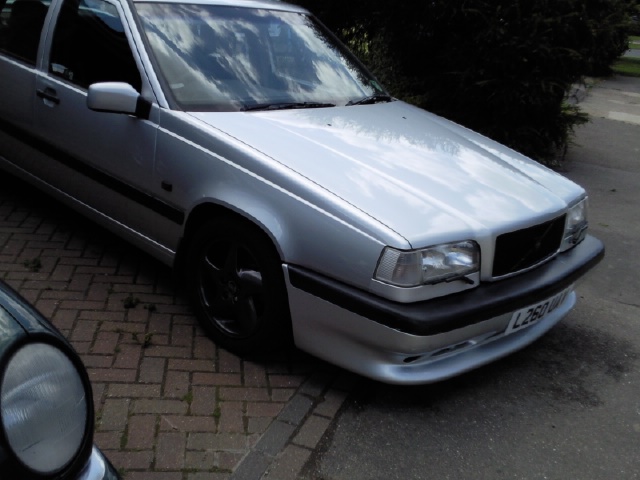 New 850 T5 Owner - Page 1 - Volvo - PistonHeads - The image shows a silver-colored car parked by the side of a road. The vehicle appears to be an old model, possibly from the 1990s or 2000s, as evidenced by its design and styling. The car is parked next to a footpath, and a brick wall is visible in the background. There is another car partially visible in the foreground, suggesting that multiple cars are parked along this stretch of road. The sky above is overcast, casting a soft light over the scene.