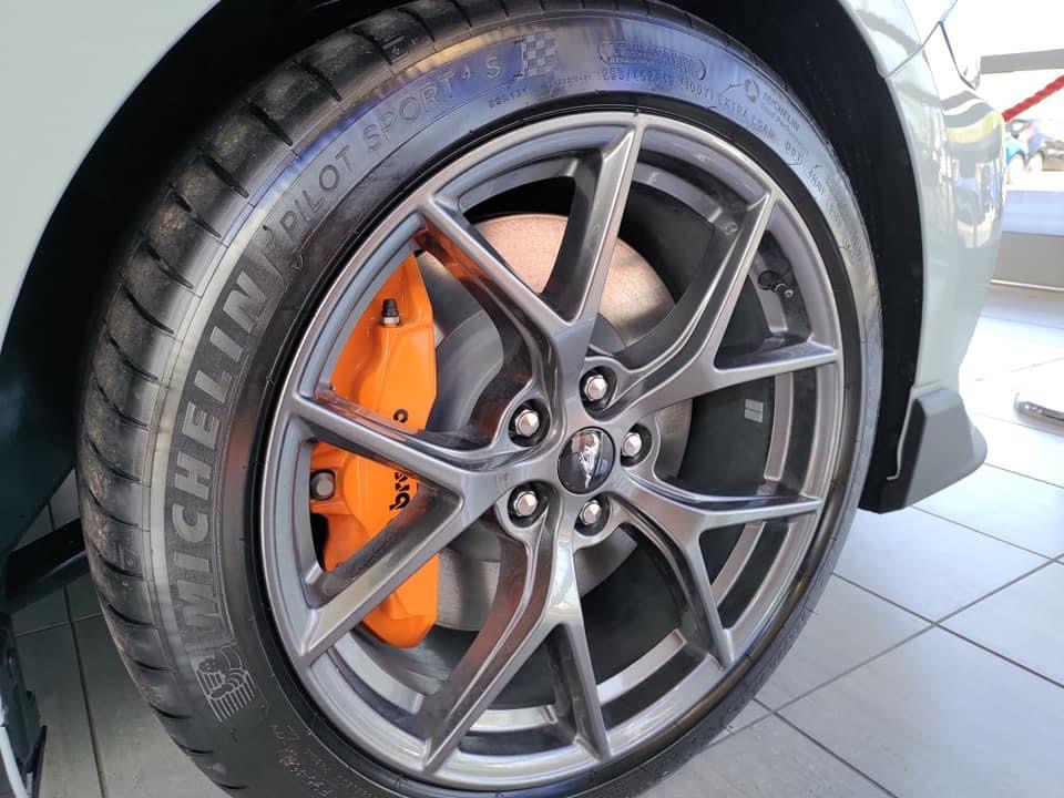 Mach 1 - Page 3 - Mustangs - PistonHeads UK - This is a photo of the front left wheel of a vehicle. The wheel has a large silver hubcap and a prominent orange brake caliper, which indicates the car's braking system is functioning correctly. The tire appears to be well-maintained with no visible damage or wear. In the background, there is a blurred image of what seems to be a person standing behind the wheel, but they are not the focus of the photo. There is also some text on the hubcap, which appears to be in a foreign language, but it's not clear enough to read. The vehicle itself isn't fully visible due to the close-up angle of the shot.