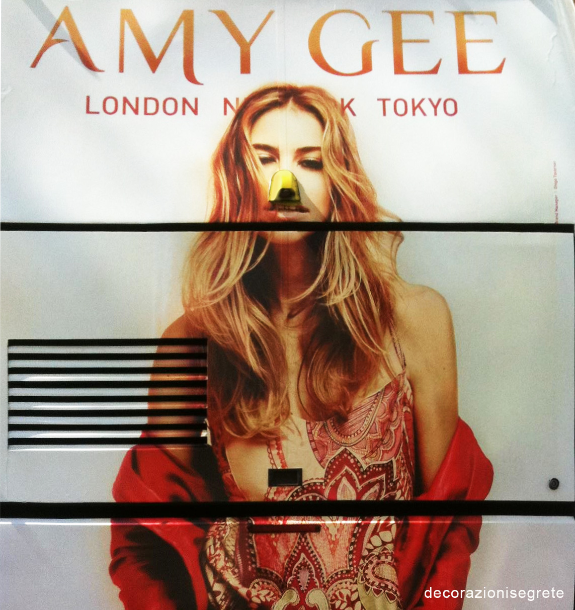 The image is a close-up photograph of a white advertisement featuring a woman. The woman, viewed from the chest up, has long blonde hair and a neutral expression on her face. She wears a red garment with a wide neckline. At the bottom of the advertisement, two metallic vents are visible, extending horizontally across the image. The background is a simple, unobtrusive light color, providing contrast to the woman's vibrant red outfit and the white of the advertisement. The text "AMY GEE | LONDON | TOKYO" is printed at the top right corner of the image in a bold, serif font.