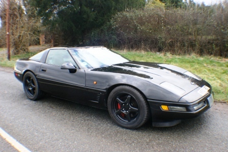 Pistonheads - The image shows a single car parked on what appears to be a suburban street. The car is a dark gray two-door coupe with a sleek and sporty design, featuring distinct creases and lines that give it a muscular appearance. The front of the car has a prominent hood, and the headlights are visible. The car is positioned at an angle to the camera, giving a clear view of its profile and the side of its hood. The setting includes scattered trees and a grassy area adjacent to the street. The road itself has visible tarmac.