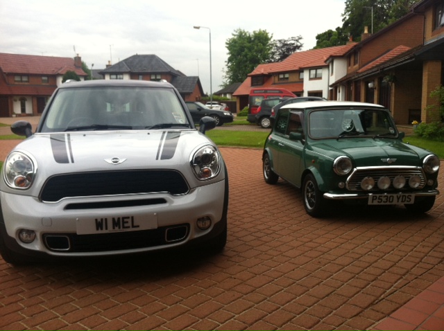 Bought a mini -  - Page 1 - Classic Minis - PistonHeads - The image shows a residential street with two classic cars parked. On the left is a silver Mini Cooper, and to its right is a green Mini Cooper. Both cars have distinguishing stripes on the hoods and are parked on a brick driveway. The vehicles are positioned in front of houses, indicating a residential setting. The sky is overcast, suggesting a gloomy weather day.