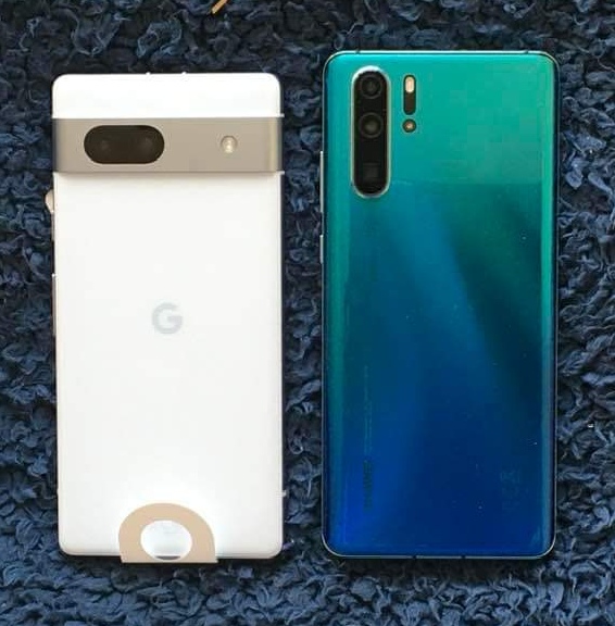 Google Pixel smartphone - Page 113 - Computers, Gadgets & Stuff - PistonHeads UK - The image displays two smartphones placed side by side on a surface. On the left is a white, rectangular smartphone with a camera module at the top center, and below it, a logo that resembles a letter "G" within a circle. On the right is a blue smartphone with a similar camera module design, also featuring a circular logo with the letters "P" inside. Both phones have fingerprint sensors below the cameras. In the background, there's a blurred image of a textured surface, possibly a wall or tabletop. The text in the image reads "MUST WATCH," indicating that this might be an instructional image about using or choosing between the two smartphones depicted.