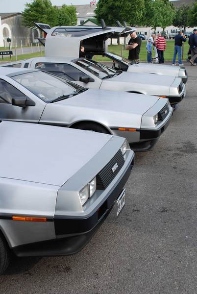 Pistonheads Supercar Sunday - The image features a row of three classic 1980s style DeLorean cars, each with an opening gull-wing design. The vehicles appear to be in excellent condition, showcasing silvertone bodies with black and gold details. They are parked on a paved surface with grass in the background. Among the cars, there are several people standing and admiring them, suggesting this might be a car show or exhibition. The setting appears to be outdoors during the daytime.